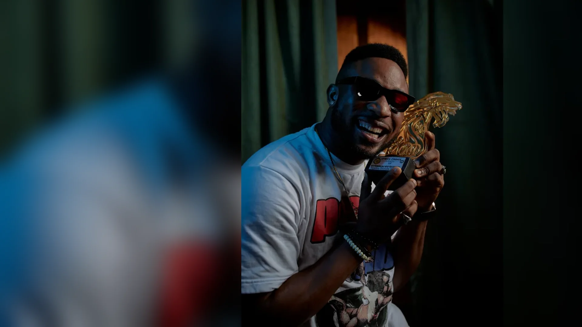 A smiling man wearing sunglasses and a graphic T-shirt holds a gold Africa Choice Awards lion trophy close to his face against a dark green curtain.