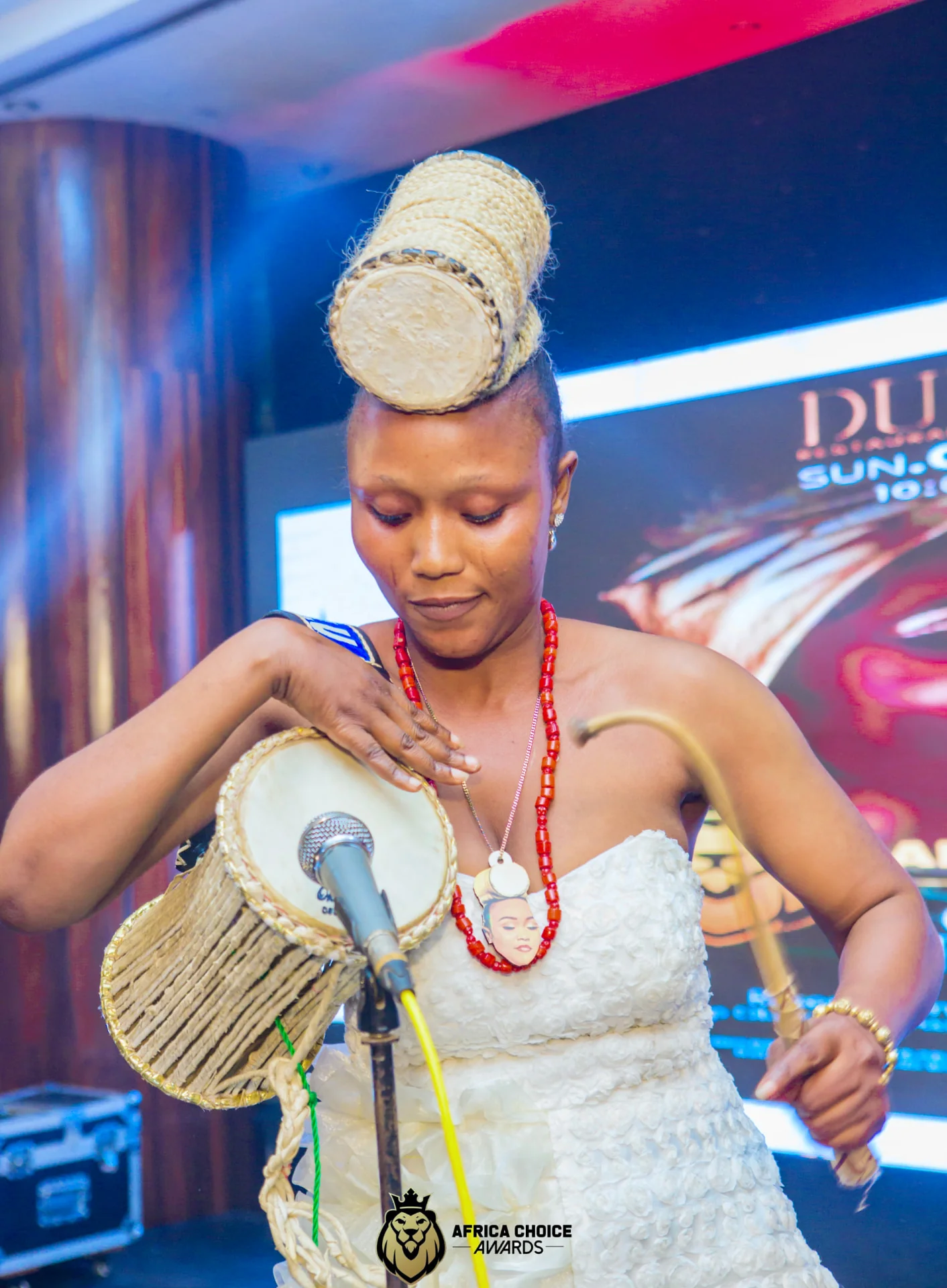 Performer in a white gown with a woven drum headpiece playing a talking drum at the microphone on the Africa Choice Awards stage