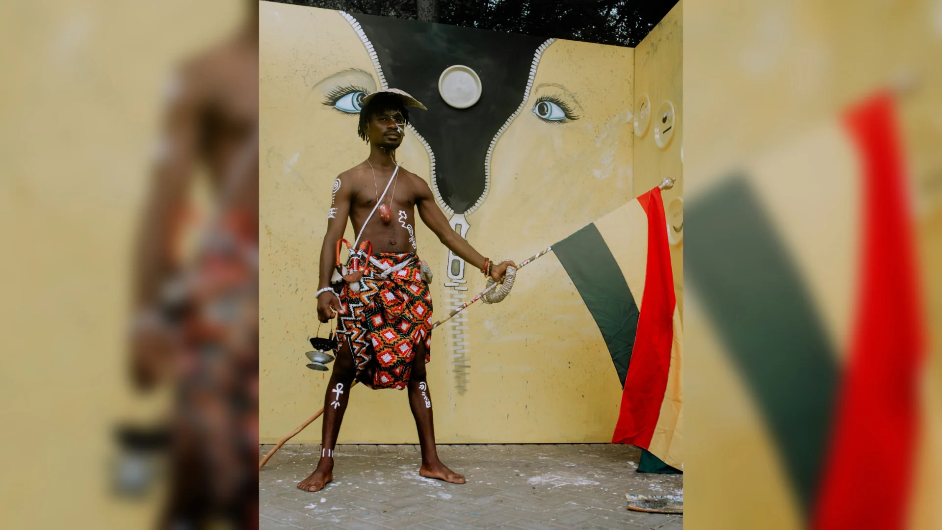 A performer in traditional African dress, painted markings, beads and a clay lamp in one hand raises a yellow, green and red striped flag in front of a painted yellow stage backdrop; centred in a wide banner with softly blurred extensions